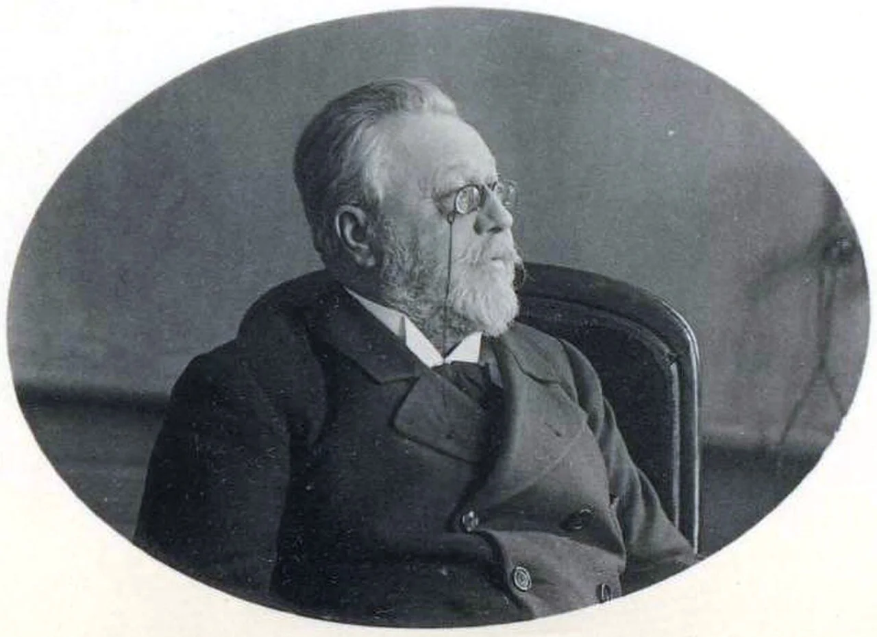 An oval-framed black-and-white portrait photograph of an elderly man with white hair and a white beard, wearing round pince-nez spectacles and a dark suit, seated in a chair and gazing pensively upward.