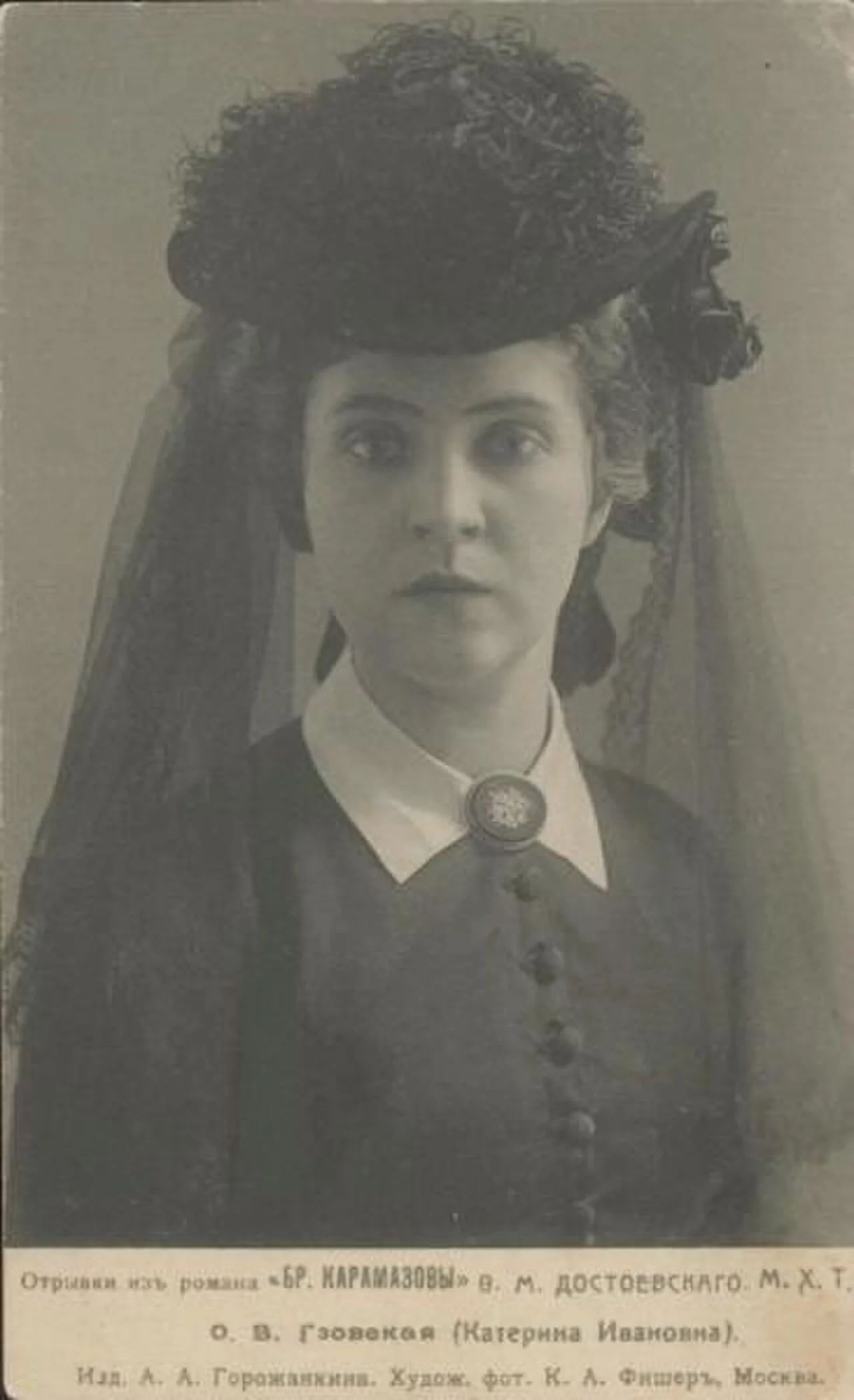A black-and-white portrait photograph of a young woman in Victorian-era theatrical costume, wearing a large ornate black hat with veil and a dark buttoned dress with white collar and brooch, gazing directly at the camera with a composed, serious expression. Depicts: Katerina Ivanovna.