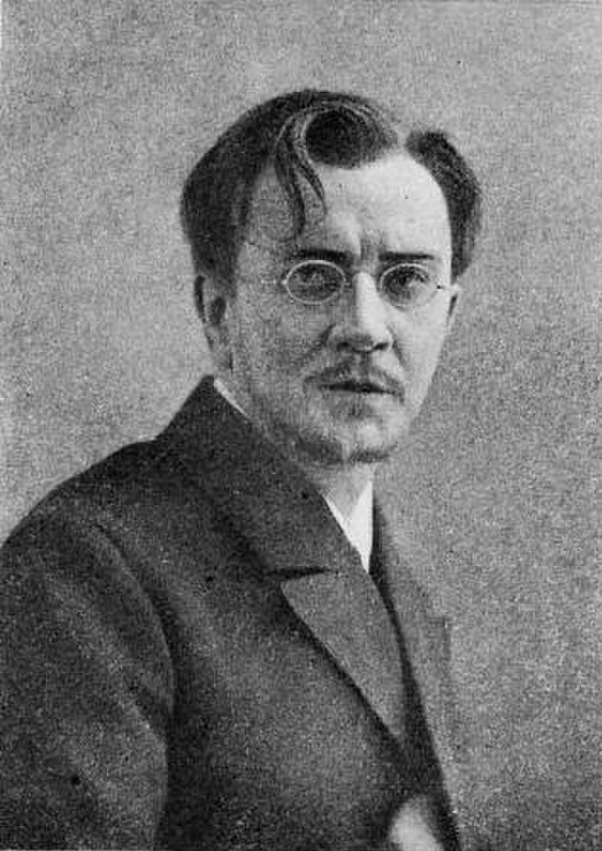 A black-and-white portrait photograph of a young man in a dark suit and round wire-rimmed glasses, gazing intently at the camera with a serious, brooding expression. Depicts: Ivan Karamazov.