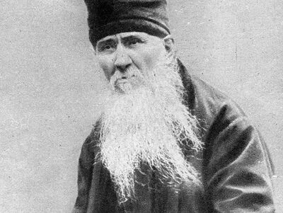 Black-and-white photograph of an elder with a vast white beard and black skullcap — the Optina-elder prototype behind Zosima. Depicts: Elder Zosima.