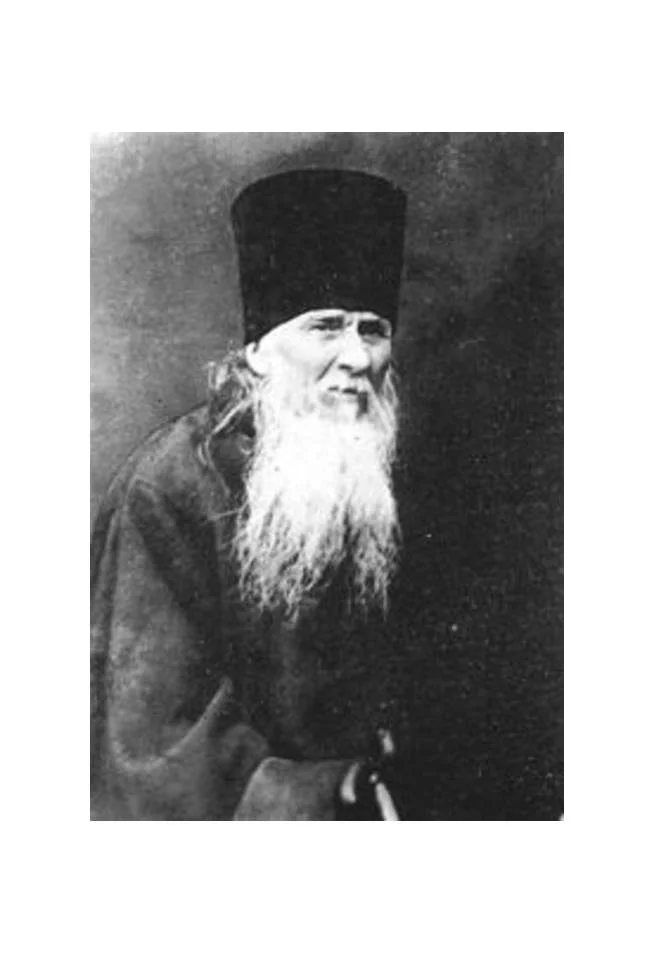 A black-and-white photograph of an elderly bearded man wearing a tall black Orthodox monastic hat (klobuk), with a long white beard, dressed in dark robes, gazing slightly upward. Depicts: Elder Zosima.