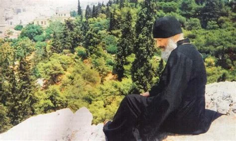 An elderly man in black Orthodox Christian monastic robes sits contemplatively on a rocky outcrop overlooking a green, tree-filled hillside landscape.
