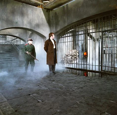 A dimly lit prison or dungeon interior with stone arches and iron bars, where a soldier holding a rifle stands guard over a tall man in a long dark coat amid drifting smoke. Depicts: Dmitri Karamazov.