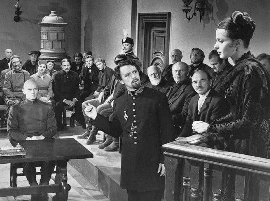 Film still: the trial — the defendant in uniform gestures from the dock before a packed courtroom. Depicts: Dmitri (Mitya) Karamazov.